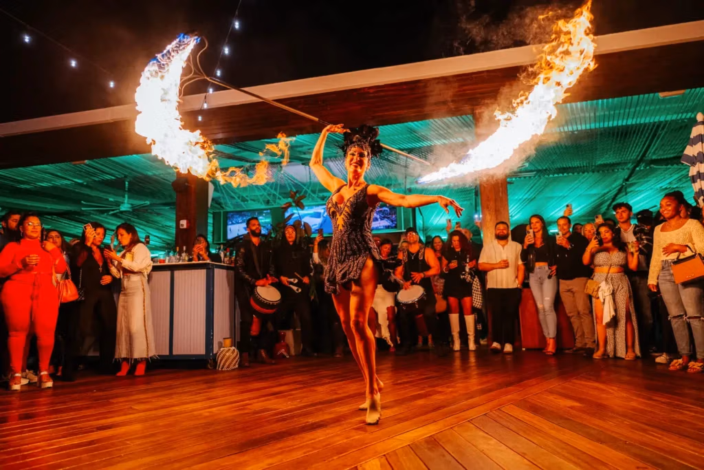 Fire performer spins two flaming staffs in a dark indoor venue as a cheering crowd watches from a wooden floor.